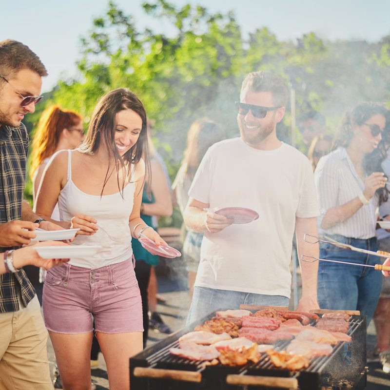 Group of people standing around grill, chatting, drinking and ea