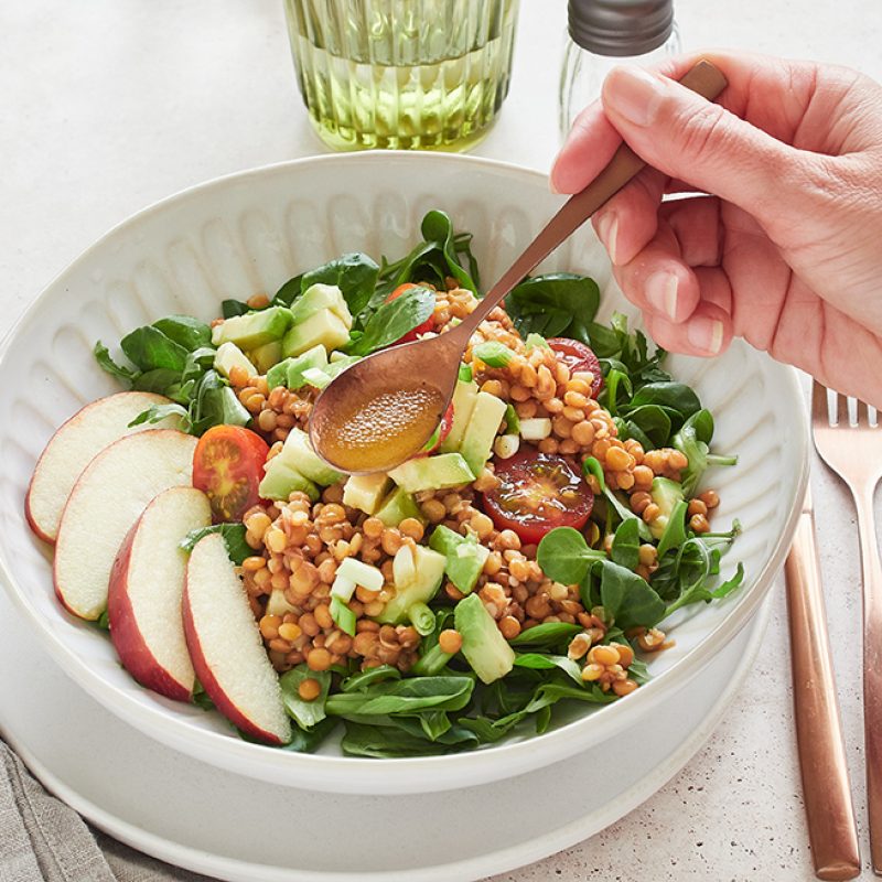 Close-up view: hand holding spoon with vinaigrette dressing salad of greens, lentils, avocado and apple. Healthy food.
